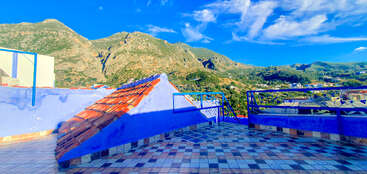 Une terrasse carrelée aux murs bleus surplombe des montagnes verdoyantes sous un ciel radieux. Des balustrades en fer forgé et des tuiles rouges ajoutent un charme méditerranéen.