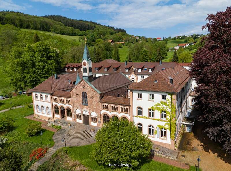 Cette image montre un bâtiment historique pittoresque doté d'une tour horloge centrale, entouré d'une végétation luxuriante, d'arbres et de collines, sous un ciel bleu partiellement nuageux.