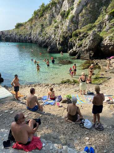 Des gens se détendent sur une plage rocheuse au bord d'une eau bleue et limpide, entourée de falaises. Certains prennent le soleil sur des serviettes, tandis que d'autres nagent ou pataugent dans la mer.
