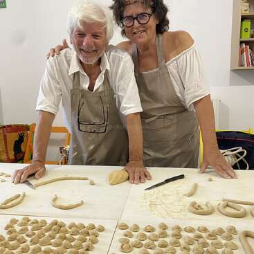 Una pareja de ancianos sonríe en la cocina, con delantal. La masa y los trozos de pasta casera están esparcidos por la mesa espolvoreada de harina que tienen delante.