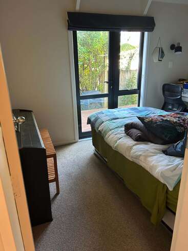 A cozy bedroom with a neatly made bed, green bedspread, pillows, desk with chair, dresser, small bench, and glass doors opening to a garden.