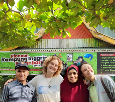 Quatro pessoas, sorrindo juntas, estão embaixo de galhos frondosos em frente a uma placa colorida "Kampung Inggris", indicando um programa de inglês. A atmosfera é alegre e amigável.