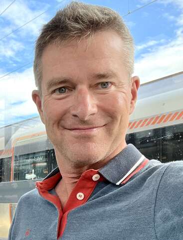 Ein lächelnder Mann macht draußen ein Selfie und trägt ein blau-rotes Hemd mit Kragen. Hinter ihm sind ein moderner Zug und ein blauer Himmel mit Wolken zu sehen.
