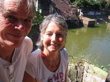 L'image représente un couple âgé posant pour un selfie devant un plan d'eau, avec un bâtiment en arrière-plan.