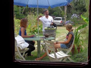 L'image montre un homme et deux femmes assis à une table dans un jardin, avec une voiture et des arbres visibles à l'arrière-plan.
