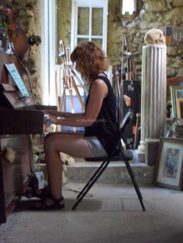 L'image représente une femme jouant du piano dans une pièce au mur de pierre, entourée de divers objets, dont un crâne sur un piédestal.