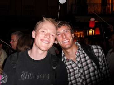 L'image représente deux hommes posant pour une photo, l'homme de gauche portant une chemise noire et celui de droite une chemise à carreaux.