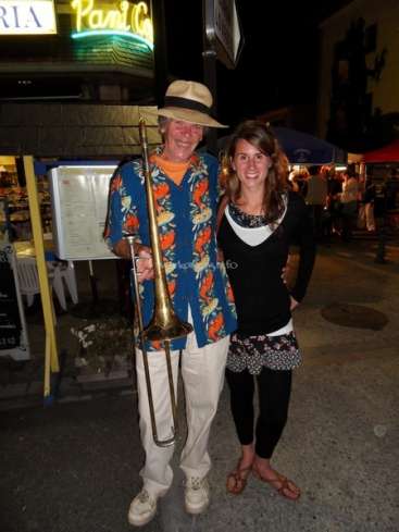 L'image représente un homme et une femme posant pour une photo dans une rue la nuit, l'homme tenant un trombone. L'homme porte une chemise et un chapeau hawaïens.