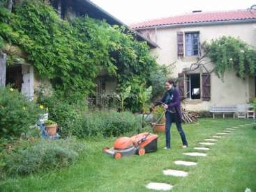 L'image représente une femme en train de tondre une pelouse devant une maison, entourée d'une végétation luxuriante et d'un sentier en pierre menant à l'entrée.