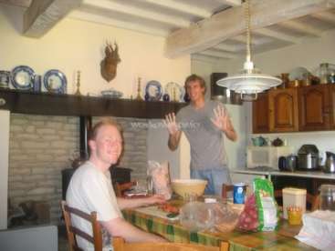 L'image représente deux hommes dans une cuisine, l'un debout et l'autre assis à une table, entourés de divers aliments et ustensiles de cuisine.