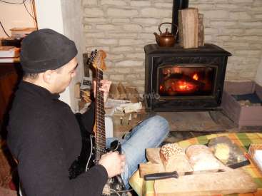 L'image représente un homme jouant de la guitare devant un poêle à bois, avec au premier plan une table sur laquelle se trouvent du pain et du fromage.