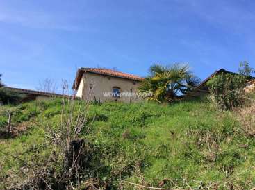 L'image représente une maison située sur une colline, entourée d'une végétation luxuriante et d'un ciel bleu clair. La maison a un toit en tuiles et une petite fenêtre.