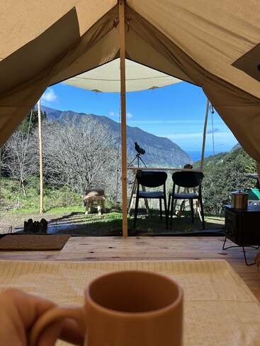 Une personne prend un café à l'intérieur d'une tente, avec vue sur les montagnes, les arbres et le ciel bleu. Des chaises, une petite table et du matériel de camping confortable créent une scène paisible en plein air.