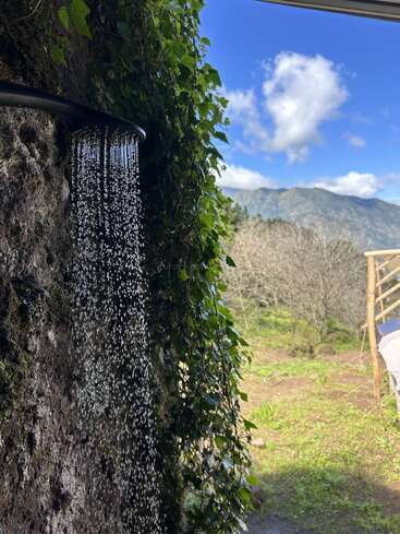 Une douche extérieure coule d'un mur rustique en pierre recouvert de lierre, avec en arrière-plan un ciel bleu vif, des montagnes et un paysage verdoyant.