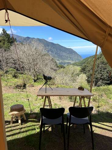 Vue d'une tente montrant une simple table en bois et deux chaises, avec des montagnes, des arbres et l'océan en arrière-plan sous un ciel bleu lumineux.