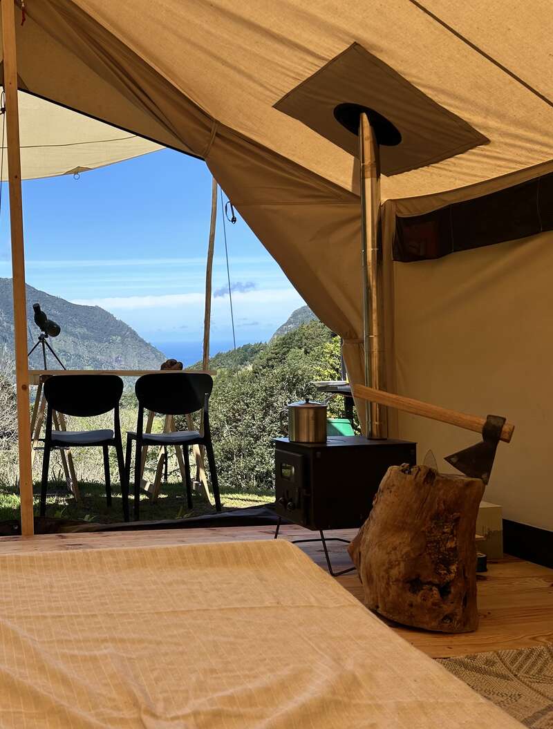 À l'intérieur d'une tente confortable, un poêle à bois, des chaises et une table offrent une vue imprenable sur les montagnes. Une hache repose sur un rondin, promettant aventure et tranquillité.