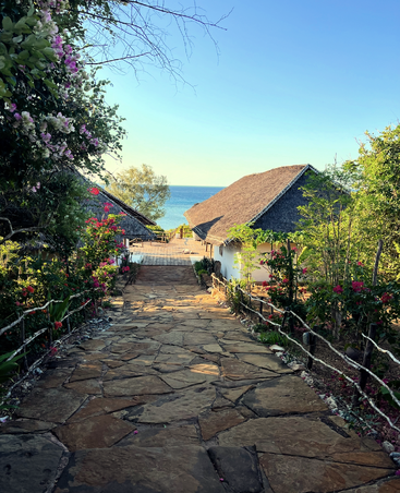 Ein Steinweg führt durch bunte Blumen und üppiges Grün zu charmanten Strohdachhütten mit Blick auf das ruhige blaue Meer unter einem strahlend klaren Himmel.