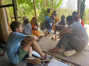 Eine bunt gemischte Gruppe von Menschen, darunter Männer und Frauen, sitzt in einem schattigen Außenbereich zusammen und unterhält sich, umgeben von Grün und natürlichem Licht.