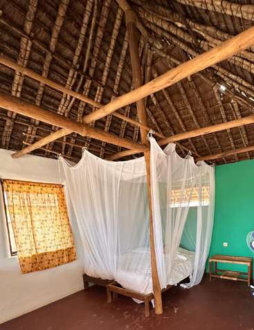 Ein rustikales Schlafzimmer mit einem Strohdach, Holzbalken, einer grünen Wand, einem Moskitonetz über dem Bett, einem geblümten Vorhang, einfachen Holzmöbeln und einem Tischventilator.