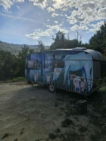 Uma caravana pintada de forma criativa fica ao ar livre, cercada por árvores e grama. O mural apresenta um cavalo branco, uma vaca e um cenário de paisagem sob um céu nublado.