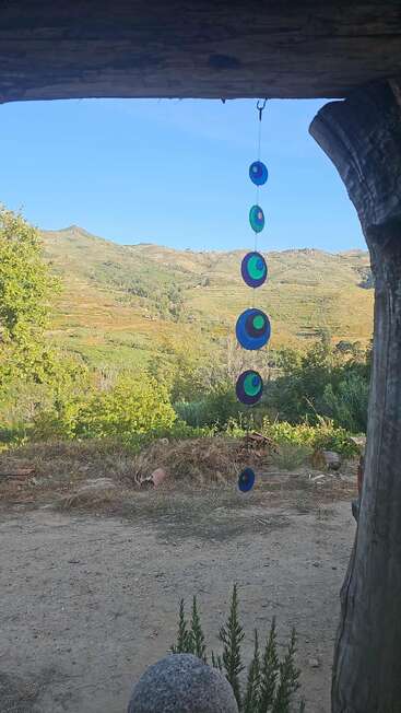 Esta imagem mostra uma vista através de uma moldura de madeira rústica em direção a colinas verdes, com uma decoração colorida pendurada em primeiro plano e um céu azul claro acima.