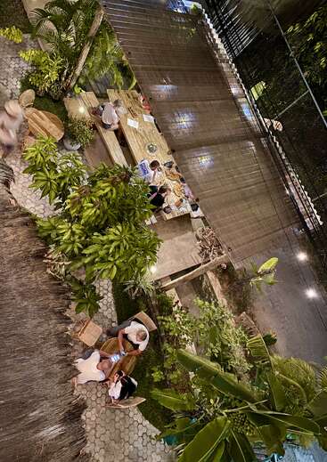 The image depicts a serene outdoor dining area, featuring a long wooden table surrounded by lush greenery and a few people enjoying their meals.