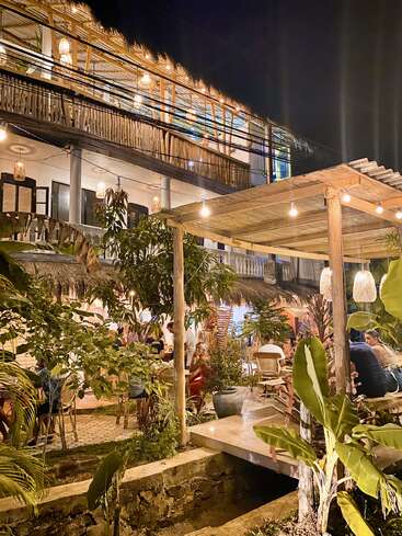 The image depicts a tropical restaurant at night, featuring a two-story building with a balcony and a patio area with tables, chairs, and string lights.