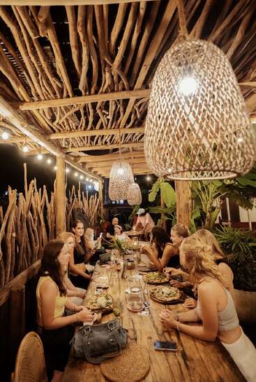 A group of people enjoy dinner together at a rustic, outdoor restaurant with wooden decor, woven lamps, and greenery, creating a warm, cozy, festive atmosphere.