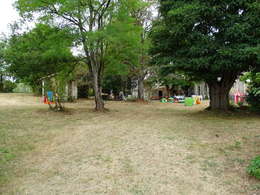 A grassy yard with large trees, children’s play equipment, and a few people in the background near a house. Colorful toys and furniture are scattered around. Relaxed atmosphere.