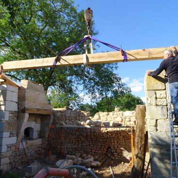 Une poutre en bois est soulevée par une grue et posée sur des murs en pierre pendant la construction. Une personne se tient sur une échelle et guide la mise en place sous un ciel dégagé.