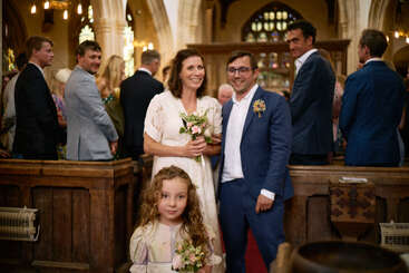 A bride and groom stand smiling in a church, holding flowers, with a flower girl in front. Guests dressed formally are seated, watching the couple.