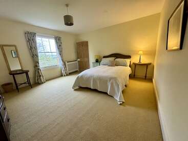 This is a spacious, cozy bedroom with a double bed, bedside tables, lamps, window with curtains, a standing mirror, and neutral-toned walls and carpet.