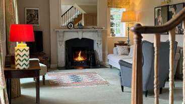 This cozy living room features a lit fireplace, colorful rug, comfortable seating, two lamps, artwork on the walls, and a staircase visible in the background.