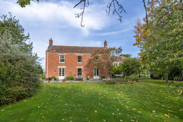 A charming two-story red brick house with large windows stands amidst a lush, green garden. Trees, shrubs, and fallen leaves surround the peaceful setting.