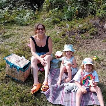 A woman and two children enjoy a picnic outdoors. They sit on a blanket with a basket, surrounded by grass and ferns, smiling and relaxed.