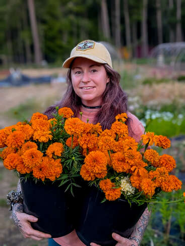 A woman wearing a beige cap stands in a garden, smiling and holding two large black buckets overflowing with vibrant orange marigold flowers, surrounded by greenery.