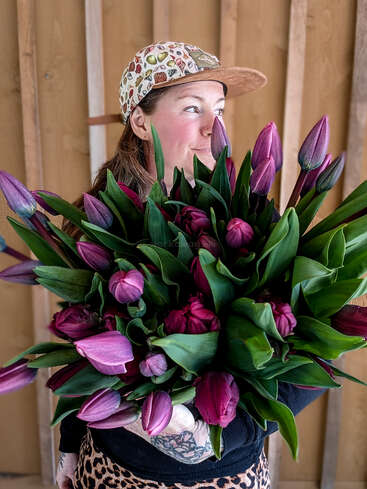 A smiling woman in a patterned cap and leopard print holds a large vibrant bouquet of deep purple tulips, standing against a wooden background indoors.