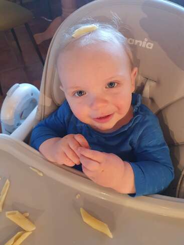 The image shows a baby sitting in a high chair with a blue shirt, surrounded by pasta, and a piece of pasta on their head, looking at the camera.