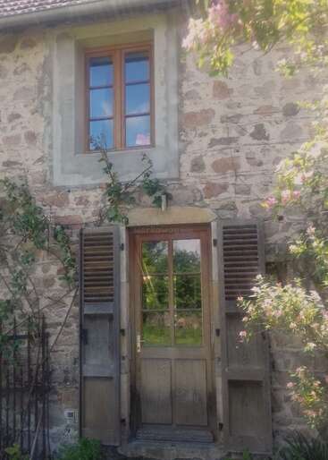 The image depicts a rustic stone house with a wooden door and window, surrounded by lush greenery and vibrant flowers, exuding a sense of serenity and natural beauty.