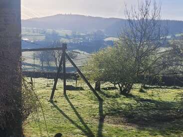 Morning sunlight casts long shadows over a grassy garden with an old wooden swing frame, leafy trees, and rolling hills in the misty distance. Peaceful countryside.