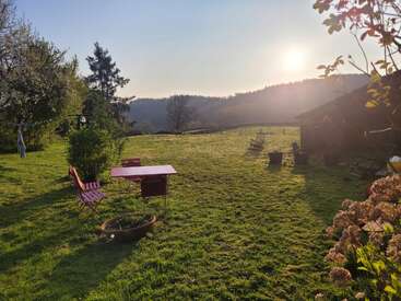 A peaceful garden scene at sunrise, featuring a red table and chairs, lush green grass, trees, distant hills, potted plants, and blooming flowers.