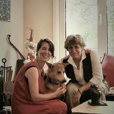 Zwei Frauen sitzen zusammen in einem Haus mit einem braunen Hund zwischen ihnen. Sie lächeln warmherzig. Der Raum ist gemütlich, mit einem großen Fenster, Kunst und Dekoration.