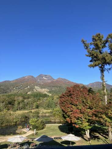 Um céu azul claro se estende sobre uma paisagem montanhosa pitoresca, com árvores verdes e em tons de outono, um amplo gramado, pessoas caminhando e vários guarda-sóis brancos ao ar livre logo abaixo.