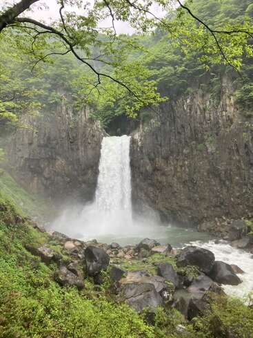 Uma cachoeira deslumbrante desce com força de um penhasco rochoso, cercada por uma vegetação exuberante e névoa, com pedras espalhadas pelo chão coberto de musgo.