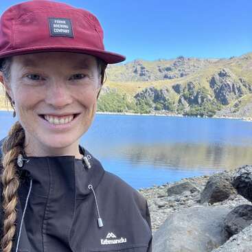 Uma mulher sorridente, usando um boné marrom e uma jaqueta preta, está à beira de um lago tranquilo, cercada por margens rochosas e montanhas verdes pitorescas, sob um céu azul claro.