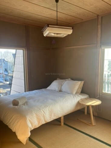 Um quarto simples em estilo japonês, bem iluminado pelo sol, com piso de tatami, uma cama baixa coberta com roupa de cama branca, dois travesseiros, uma toalha enrolada e uma pequena mesinha lateral de madeira.