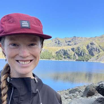 Eine lächelnde Frau mit einer kastanienbraunen Mütze und einer schwarzen Jacke steht an einem ruhigen See, umgeben von felsigen Ufern und malerischen grünen Bergen unter einem strahlend blauen Himmel.
