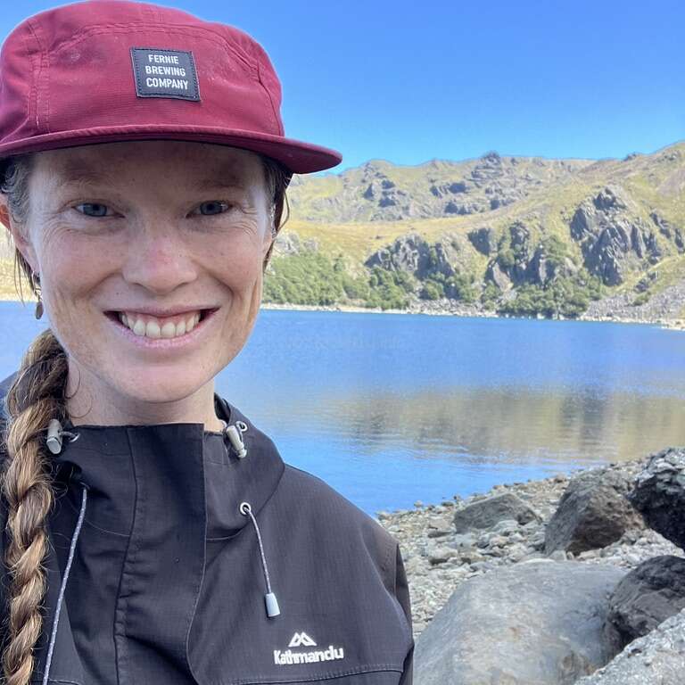 Uma mulher sorridente, usando um boné marrom e uma jaqueta preta, está à beira de um lago tranquilo, cercada por margens rochosas e montanhas verdes pitorescas, sob um céu azul claro.