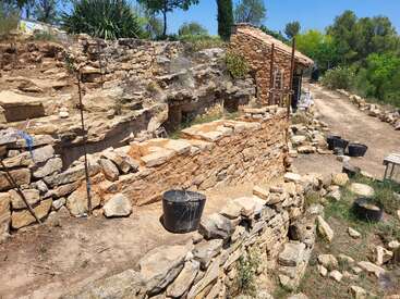 A rustic stone structure under restoration stands near a dirt path, surrounded by greenery and construction tools, buckets, and ladders on a sunny day.