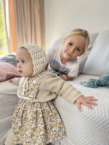 A baby in a floral dress and knitted bonnet stands by a sofa, while an older child in a white shirt smiles warmly behind her indoors.